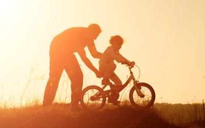 Adult helping child ride bike, sunset.