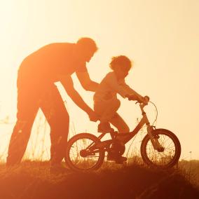 Adult helping child ride bike, sunset.
